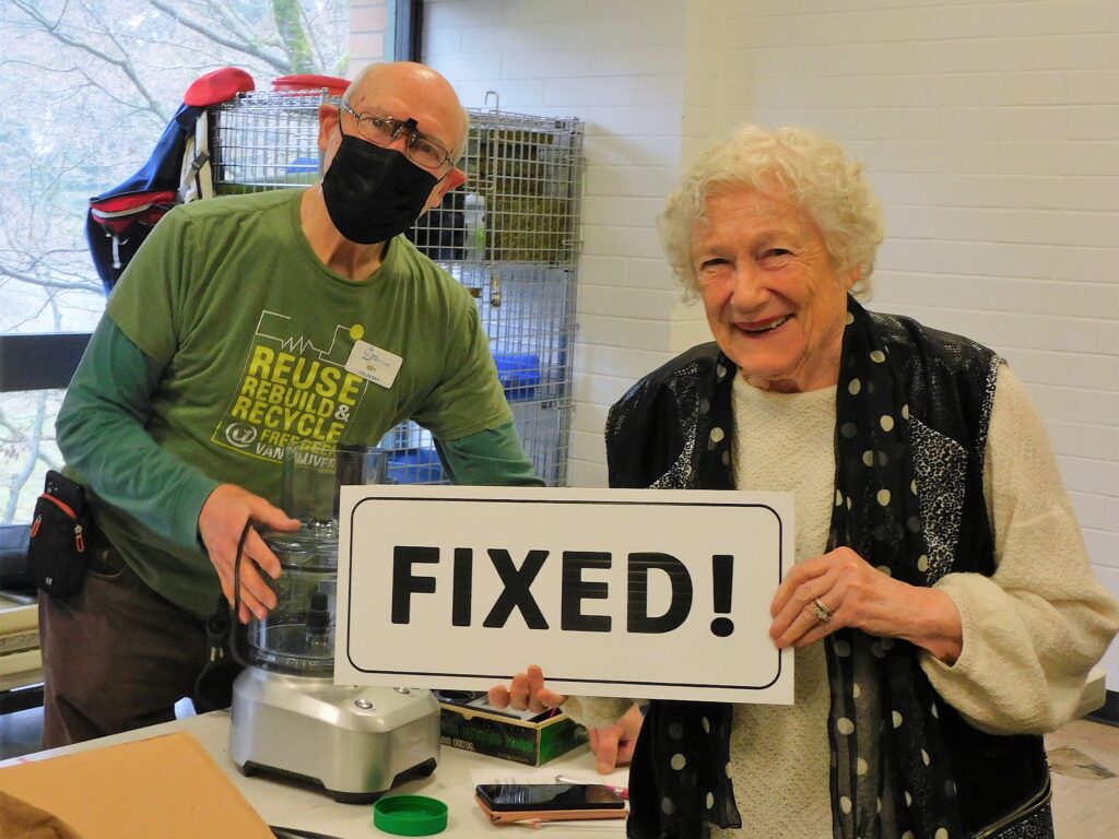 A volunteer fixer and a community member hold a "fixed" sign over a blender that was repaired at a SPEC Repair Cafe.